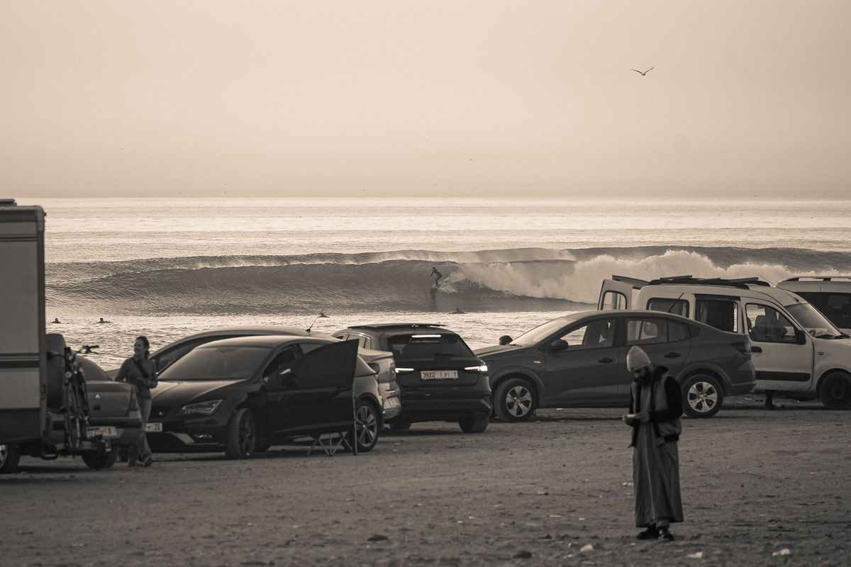 A surfer rides a long wave at Anza while people stand by parked cars on the beach road